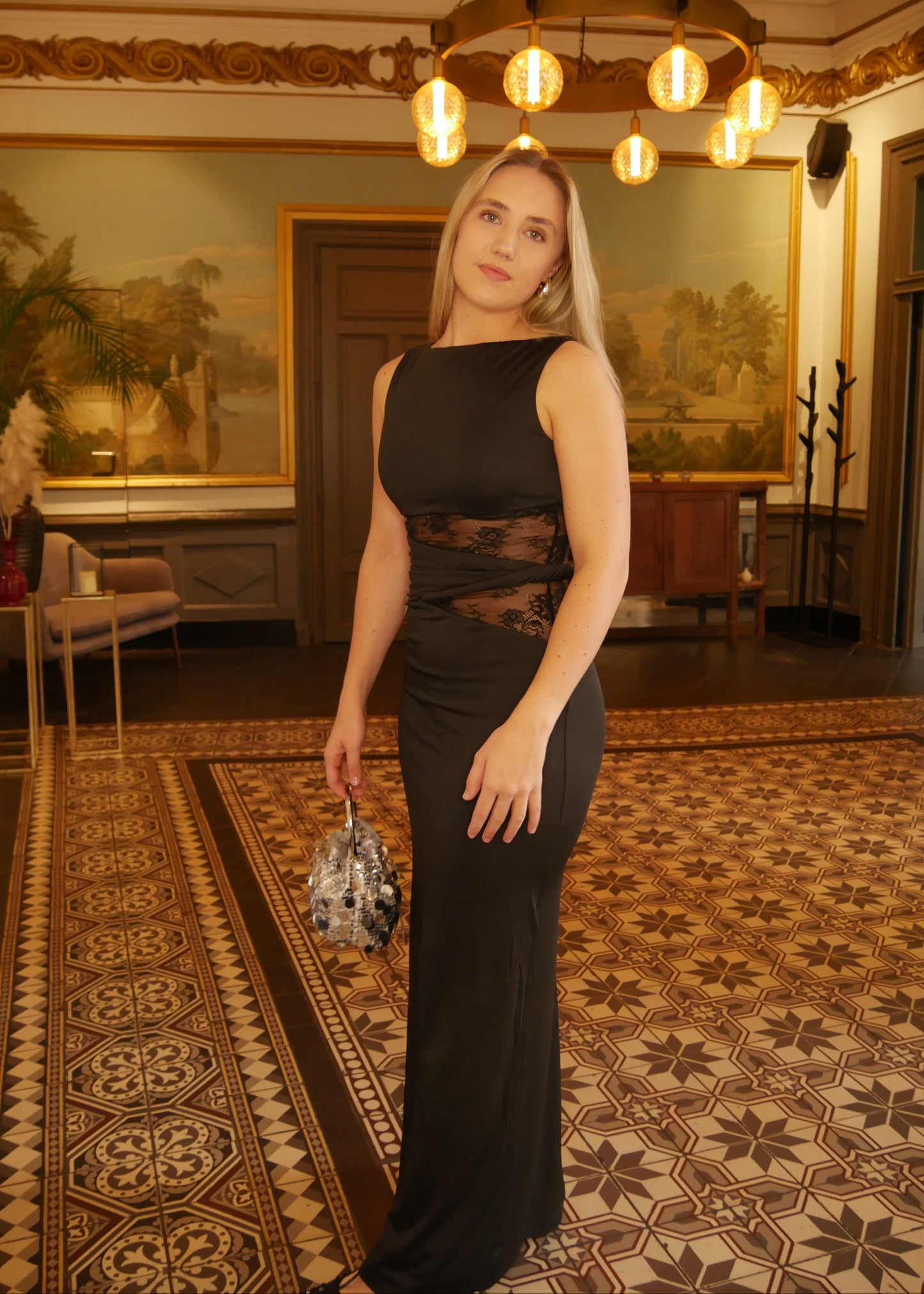 Woman in a black dress standing in an elegant room with chandelier and paintings.